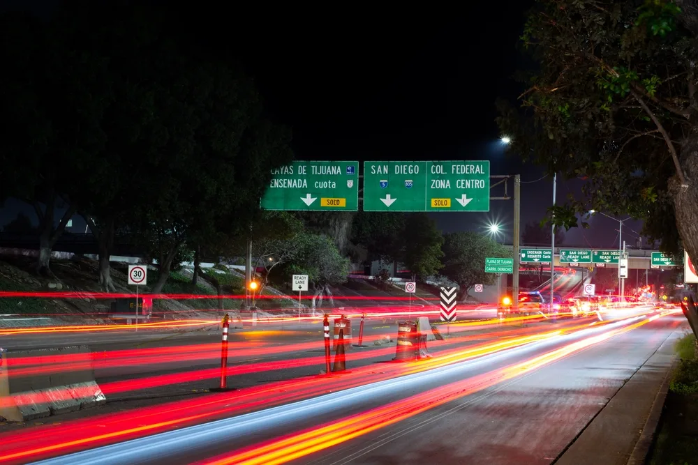 Tijuana Rio and the San Ysidro border pictured in a long exposure image with a blur of car lights making their way left to right below green street signs