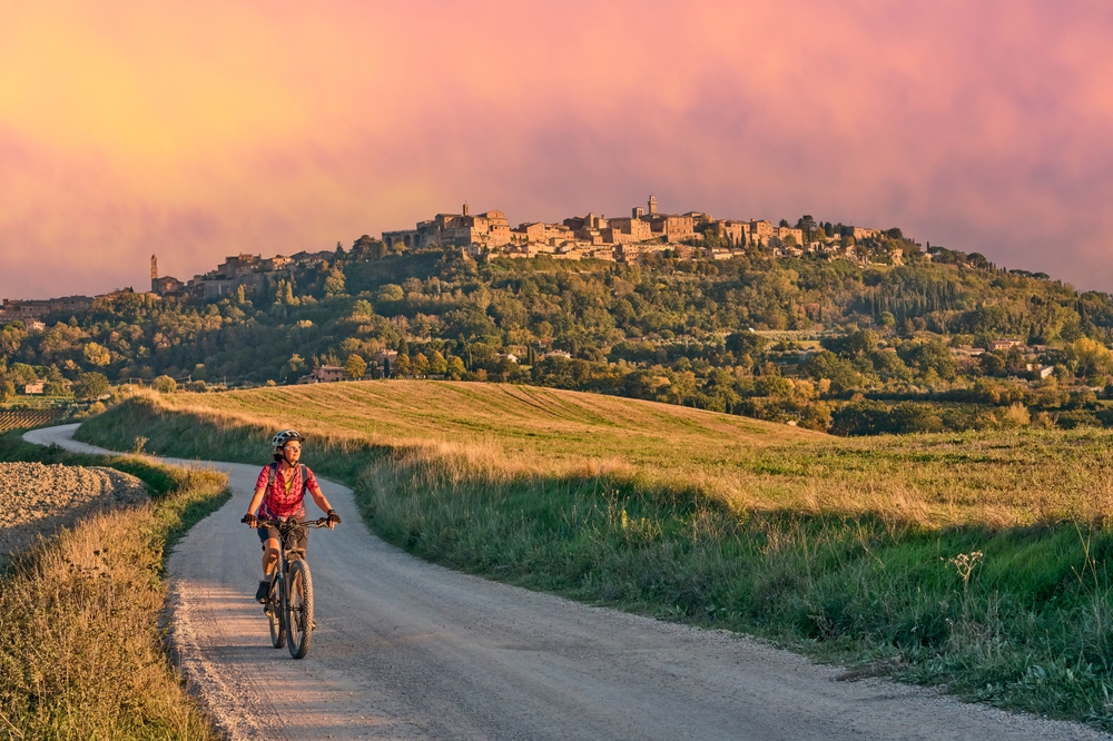 A woman riding her bike on dirt road in Ghianti with a village towering over her as an image for a piece titled Trip to Tuscany Cost