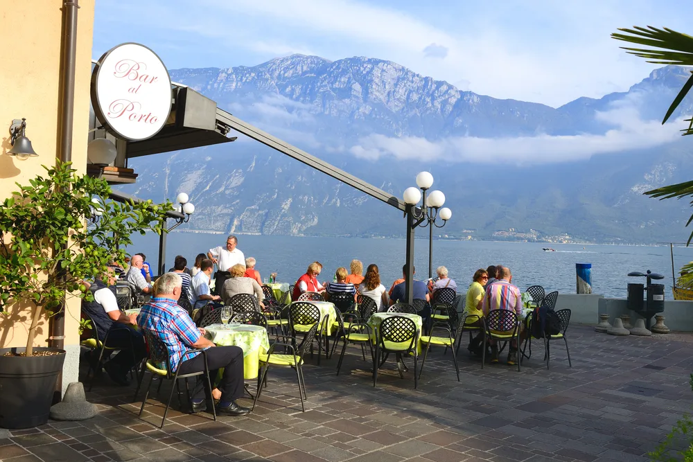 Peopl peacefully eating on a restaurant beside a lake, and a tall mountain can be seen in background. 