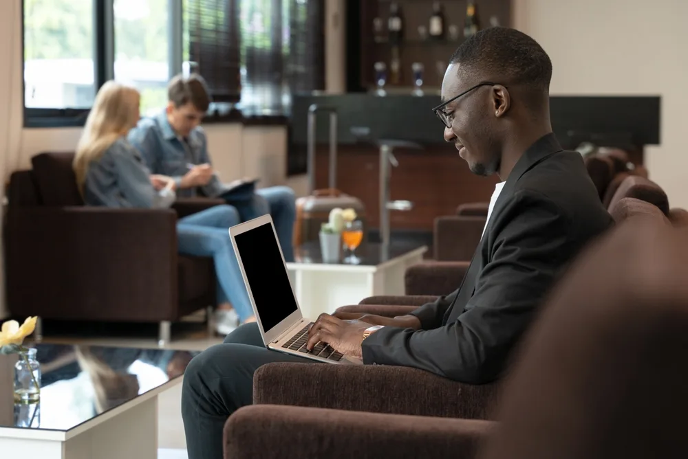 African American man smiling as he works on his laptop waiting at a Priority Pass airport lounge with others in chairs nearby