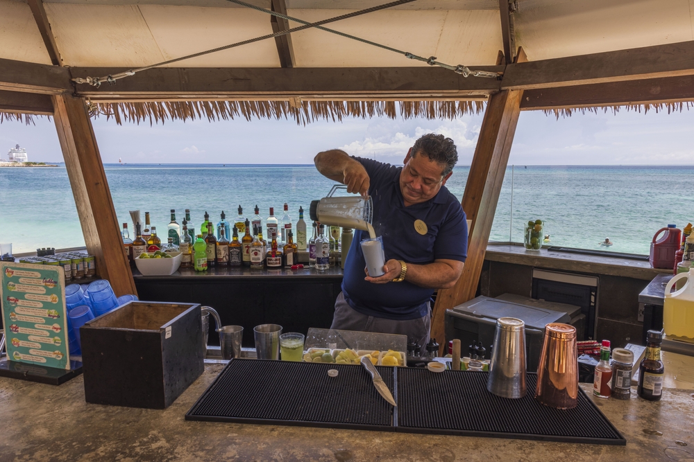 A bartender preparing a drink in a bar beside the beach, a section image on a travel guide about trip cost to Aruba.