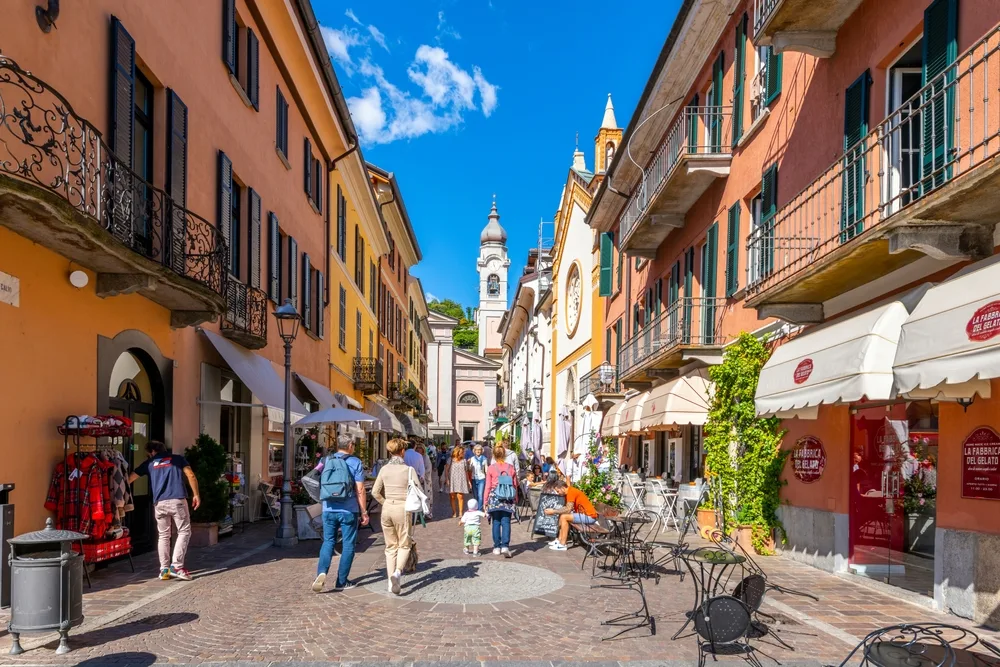 People walking on a street with shops on the ground floor of the surrounding establishments, snapped on a clear afternoon as a section image for a travel guide about safety in visiting Lake Como.