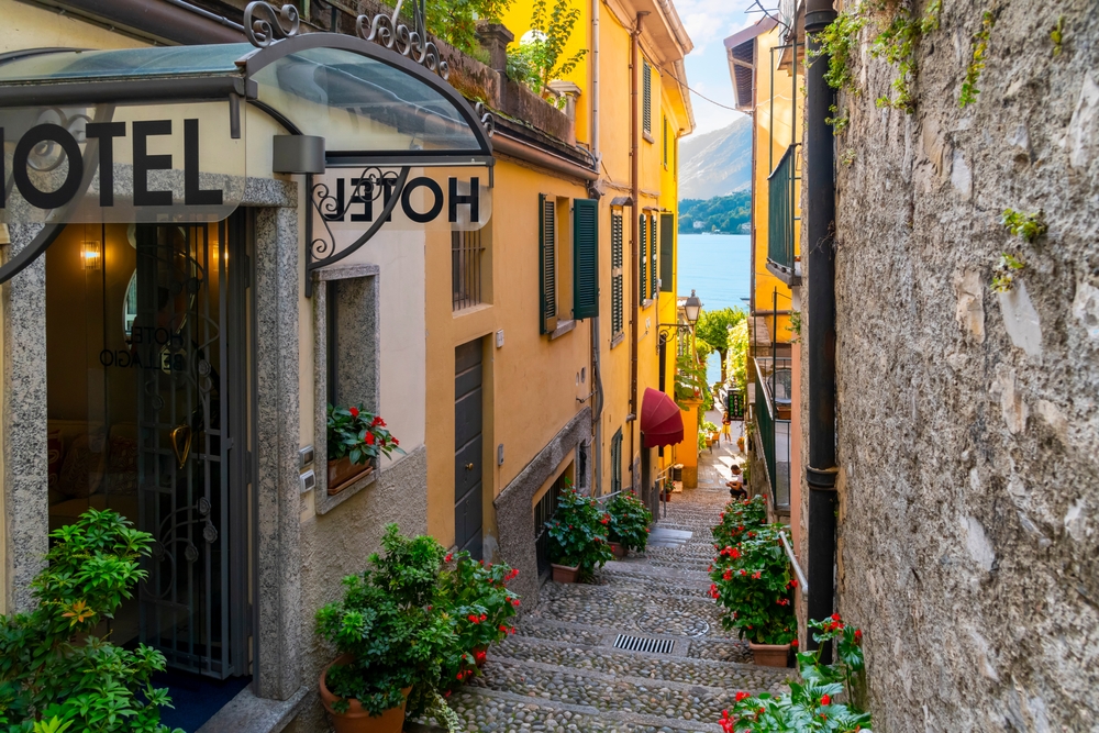 A narrow steep alley with a signage of a hotel, and a lake is visible at the end of the alley, an image for a travel guide about how safe it is to visit Lake Como.