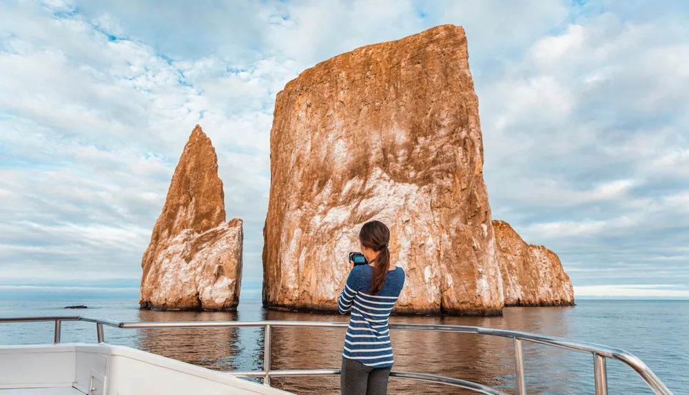 A woman standing at the edge of the boat while holding her camera and taking pictures of a unique large rocks formation at the middle of the sea. 