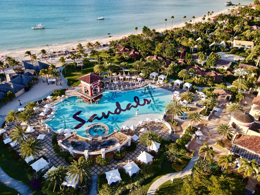 Aerial view on a resort with a large pool that writes "Sandals", and beside the resort is a calm beach with two boats. 