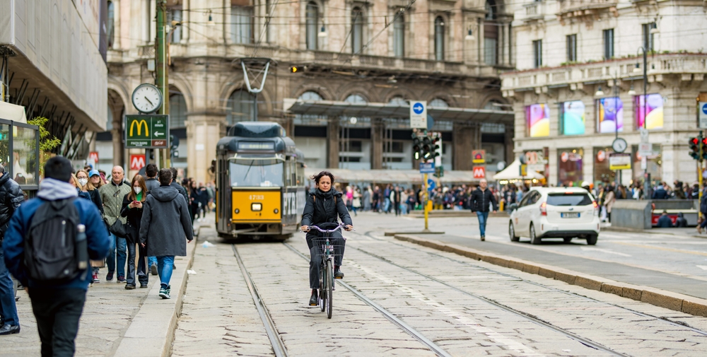 A woman can be seen biking in the middle of the trolley rails, where the trolley can be seen approaching from behind, and can be seen on the side of the trolley are people walking. 
