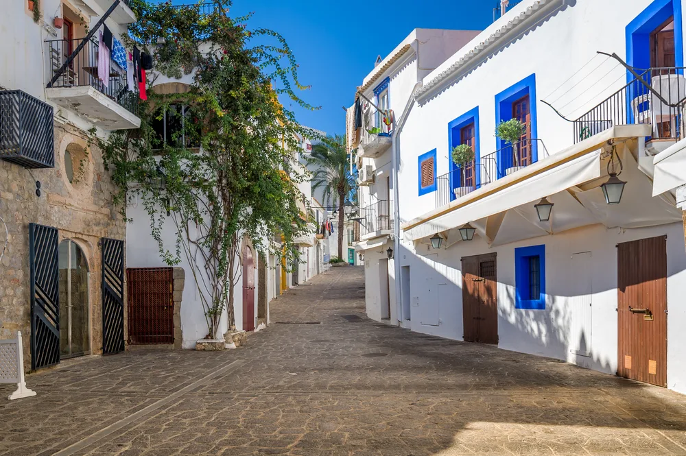Narrow pedestrian street with white buildings and blue trim pictured on a clear day for a guide to the average Ibiza trip cost
