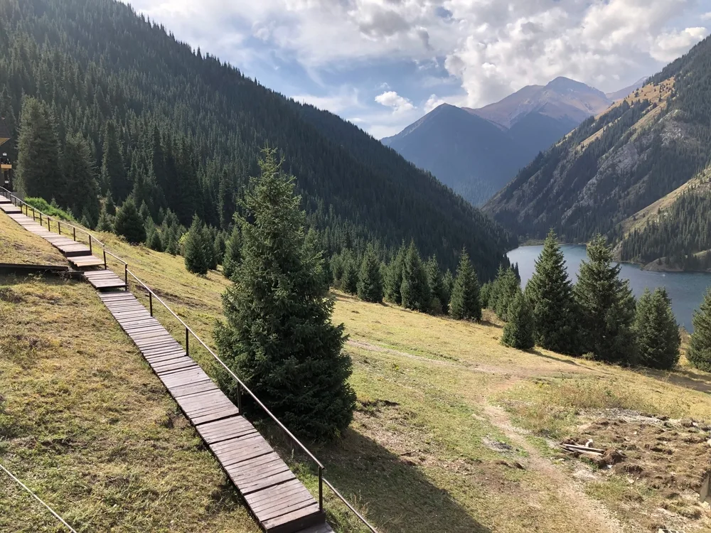 Pictured during the best time to visit Kazakhstan, the crystal-clear water in Lake Kolsai with a grassy hill located below the wooden walking path, pictured on a nice day between mountains
