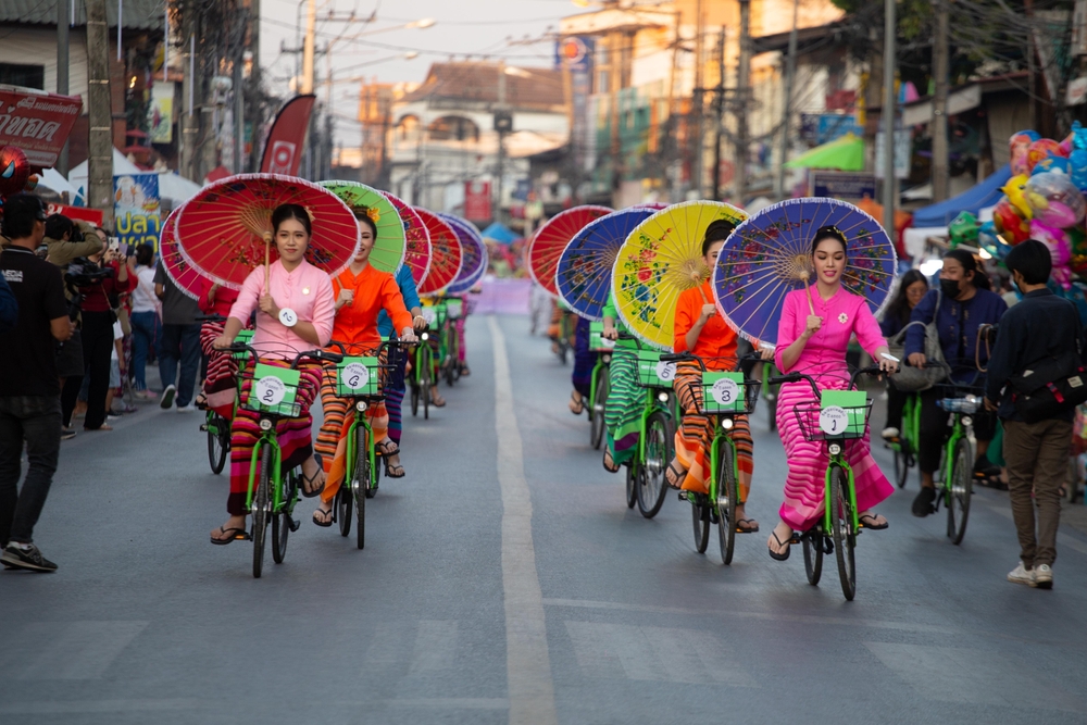 Asian women holding a vibrant traditional umbrella while riding bicycles down the street. 