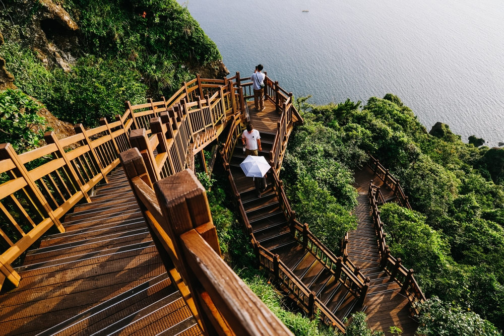 Three people walking on steep stairs at the side of the mountain, and below a calm sea can be seen, an image for a travel guide about trip cost to Korea.
