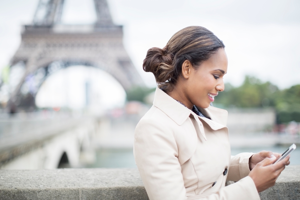 African woman uses her smartphone in front of the Eiffel Tower in Paris to show the concept of asking how much does it cost to text internationally?