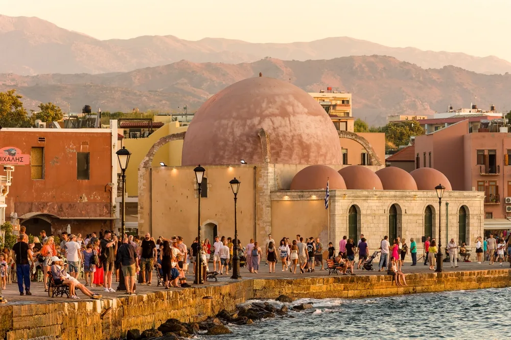 A crowded bay walk beside a mosque where people are walking and others are sitting and looking at the sunset, an image for a travel guide about safety in visiting Crete.