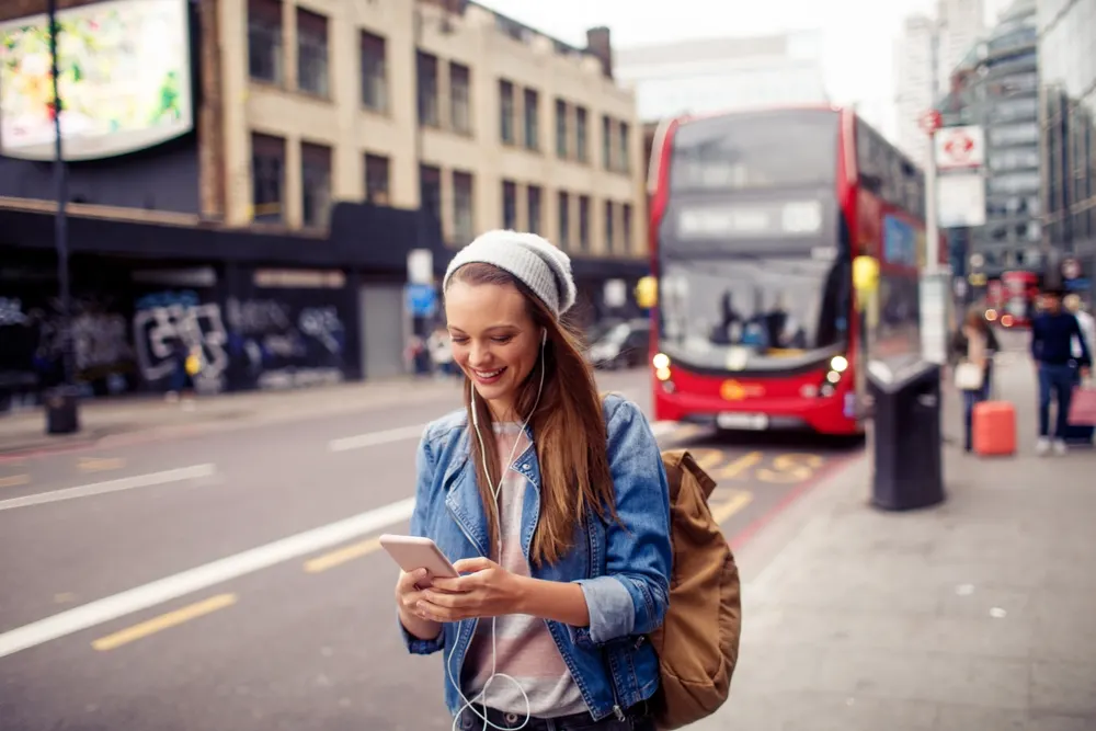 Young woman tours London with a double decker bus behind her as she uses her smartphone for a frequently asked questions section on how much does it cost to text internationally?