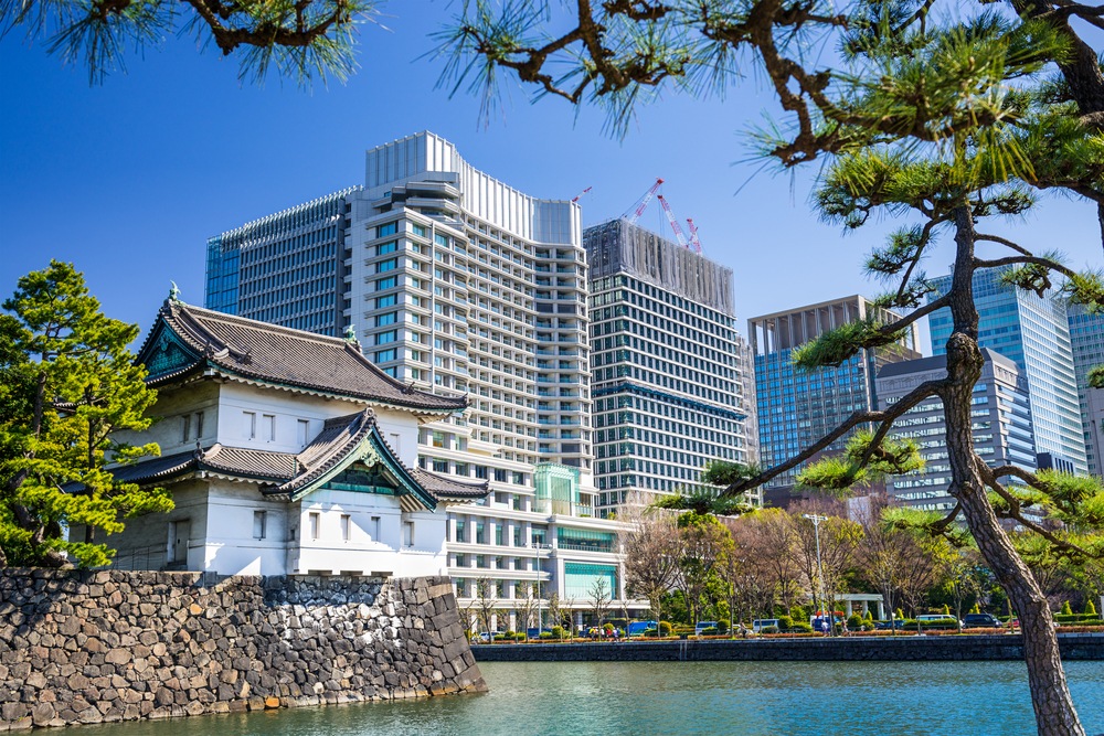 A tall hotel and a traditional structure beside a river during a clear day in Tokyo with a lake lapping the water below the scene as an image for a guide titled Trip to Tokyo Cost