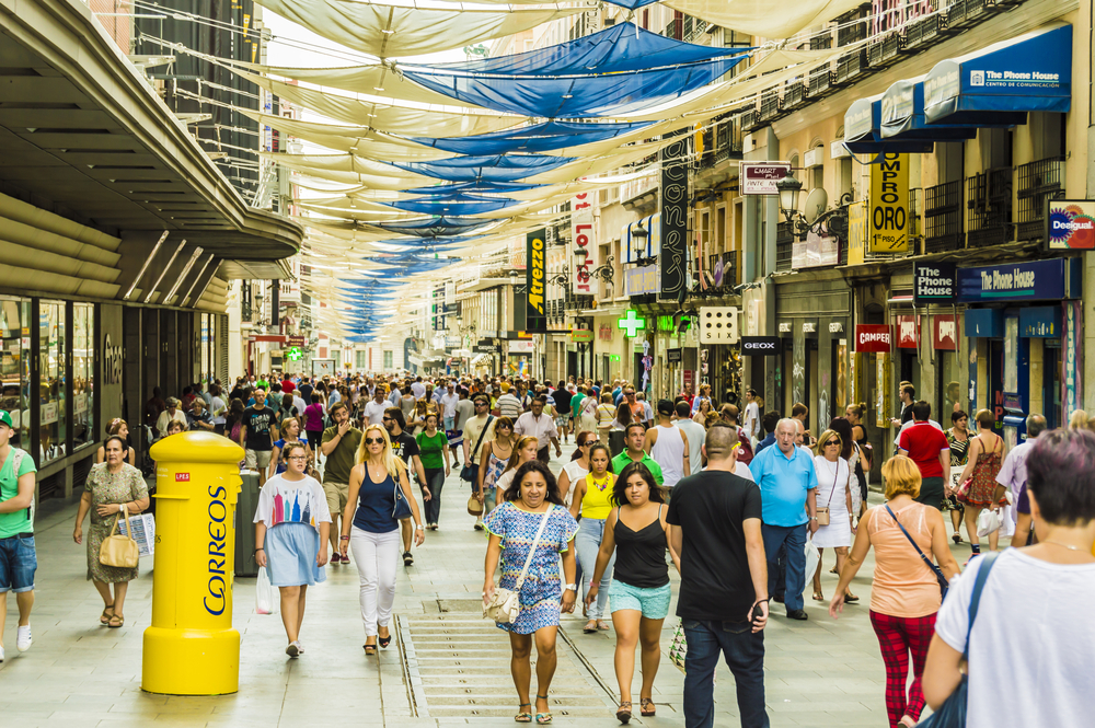 A busy street near shopping malls in Madrid where white and blue fabrics gives shade to the passing people, an image for a travel guide about safety in visiting Madrid.