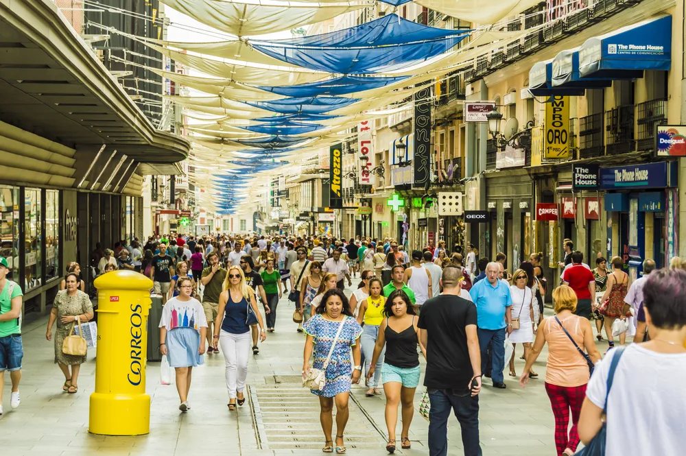 A busy street near shopping malls in Madrid where white and blue fabrics gives shade to the passing people, an image for a travel guide about safety in visiting Madrid.
