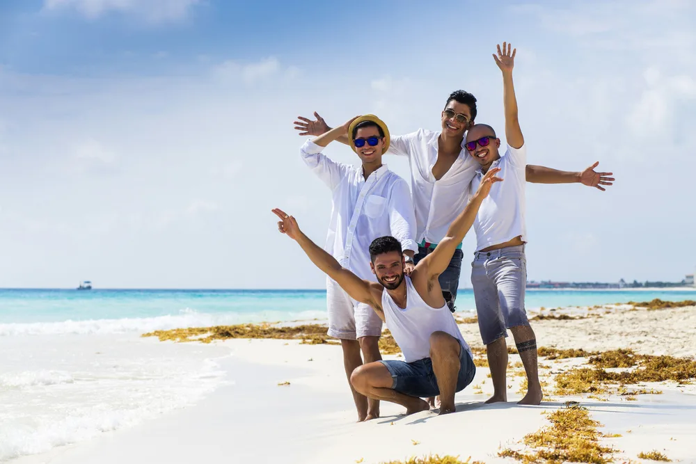 4 guy friends posing and smiling on the beach with seaweed on the white sand around them for a list of the best guys trip ideas