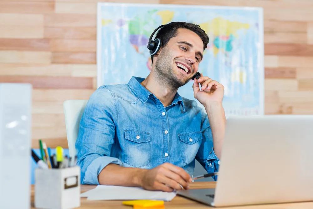 Man laughs wearing a headset while using his computer in an office with a map for a frequently asked questions section about how to become a travel agent at no cost