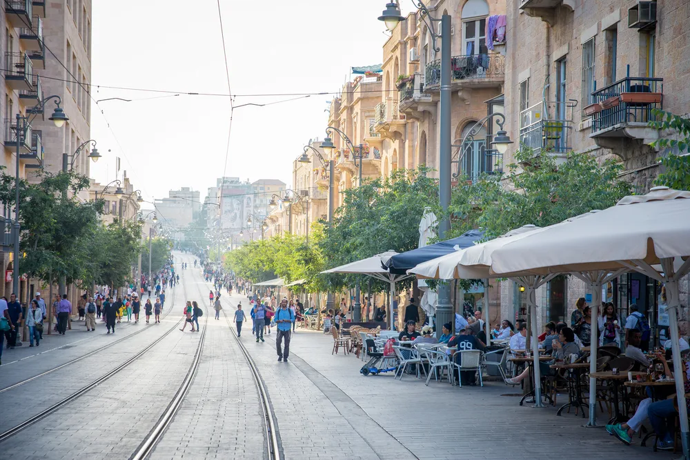 A long street with cable car rails where people are seen walking and some are sitting on outdoor restaurant chairs. 