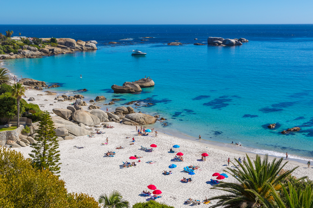 Tourists sunbathing on a beach in Camps Bay, a top pick for where to stay in Cape Town, with course looking sand, huge rocks, and clear waters. 