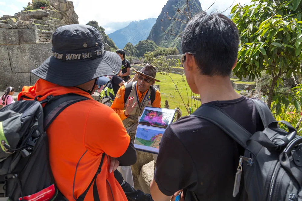 Two tourist listening to a tour guide while he explains about the place they are in using an album of printed images, a section image for an article about trip cost to Machu Picchu.