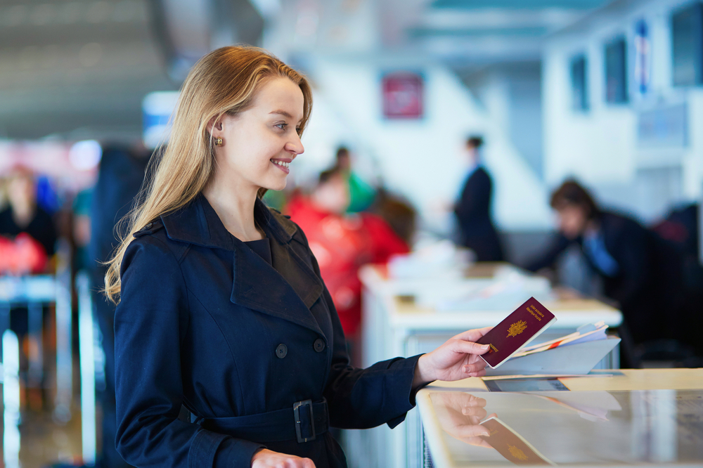 Young blonde woman provides her passport travel document number and passport book to an airport employee before traveling to a foreign country