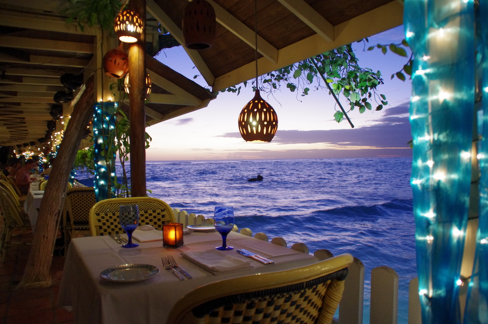 A fancy dining on the side of the ocean, where a table for two with candle at the center of the table is seen during a sunset.