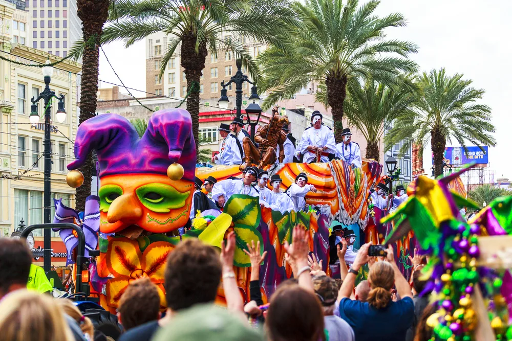 Photo of people on a Mardi Gras float with bright purple feathers and a mask making its way down the palm-lined street