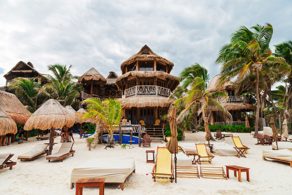 A native hotel on the beach with straw roofs and some sun beds on the shore during a overcast day. 