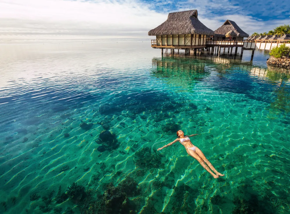 A woman wearing her two-piece swimsuit in a crystal-clear waters beside elevated native native houses in the middle of the beach, for a piece on an article about trip cost to Tahiti.