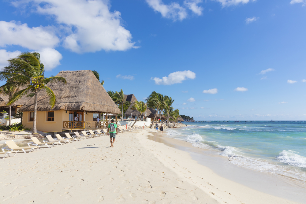 A few people walking on the fine sand of a beach with coconut trees and native huts. 