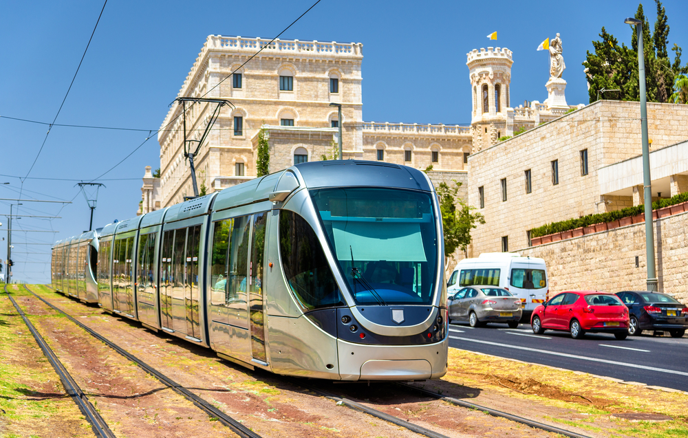 A light rail running on its tracks while cars are occupying the adjacent road, an image for a travel guide about trip cost to Jerusalem.