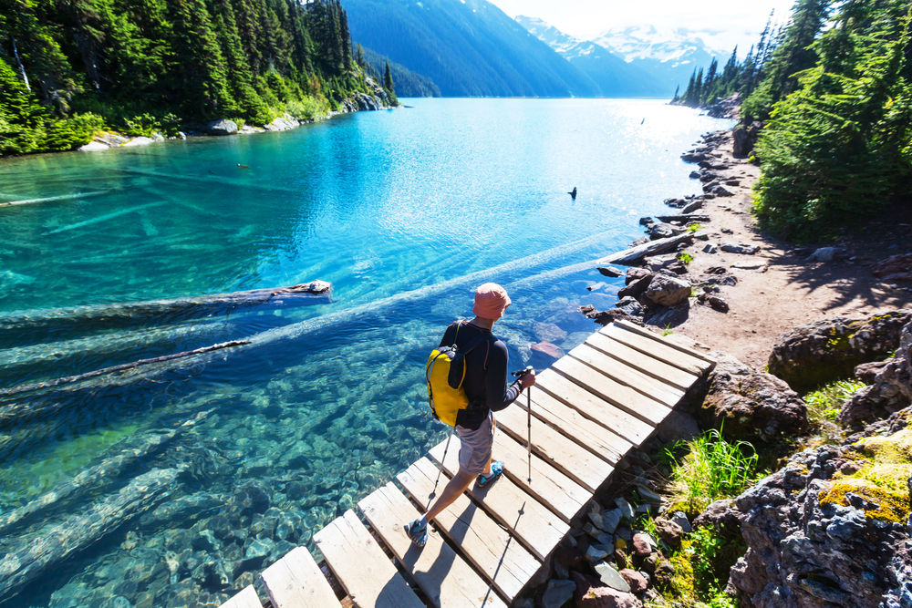 Man in shorts and a light jacket pictured hiking over a deep blue lake with clear water during the best time to visit BC