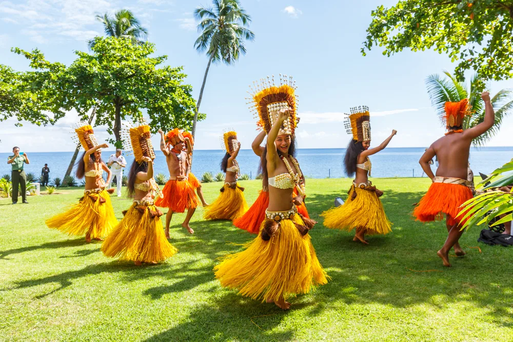 Local men and women wearing traditional tropical clothing and headdress, joyfully dancing on green grass near the sea, while a tourist is shooting a video in background. 