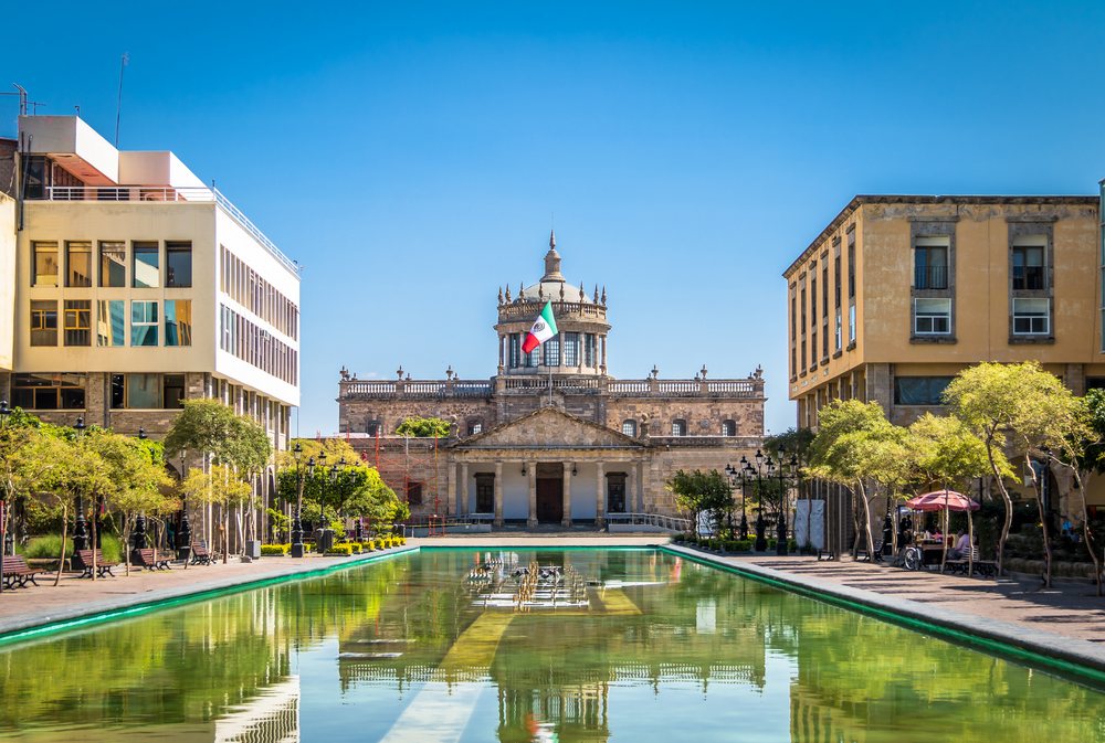 Historic old building of the Hospicio Cabanas pictured on a blue-sky day for a guide titled Is It Safe to Visit Guadalajara with its green reflecting pool between two modern buildings