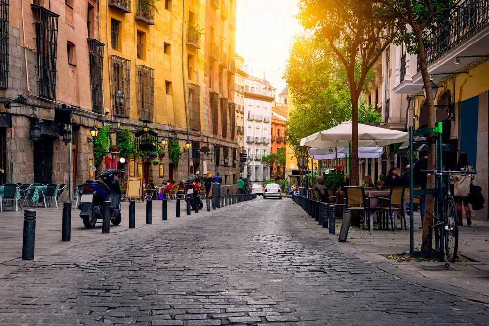 View of an old street paved with bricks, where parked motorcycles and restaurant chairs and tables and behind the metal bollards, captured during a sunset for a travel guide about safety in visiting Madrid.