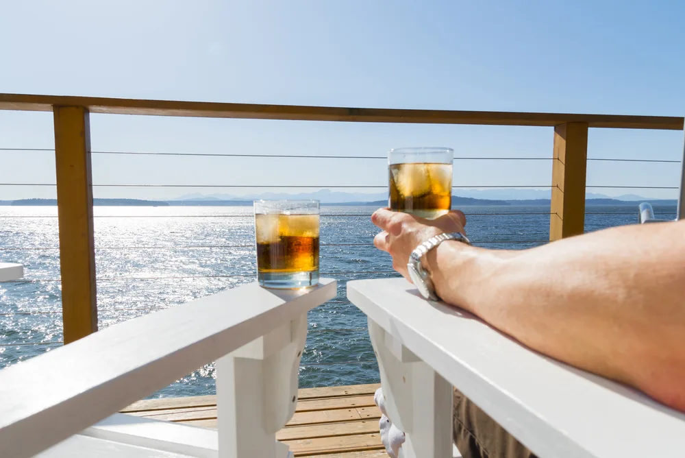 Man's arm holding a bourbon drink on a cruise ship with another empty chair next to him on the deck for a list showing the best places for guys to go for a brocation