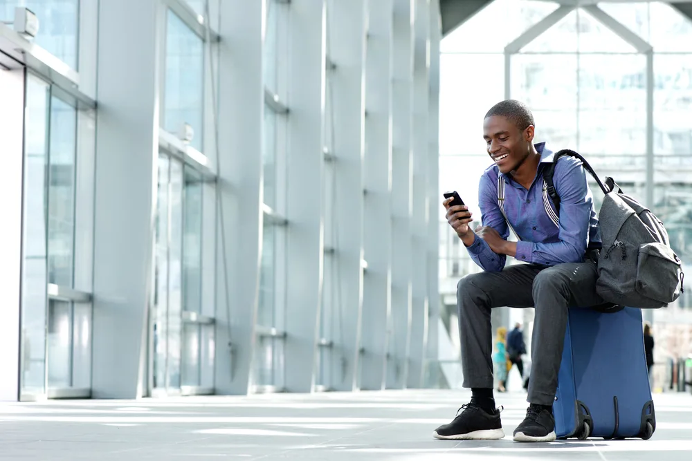 Young black businessman sits outside an airport using his smartphone to text internationally and smiling