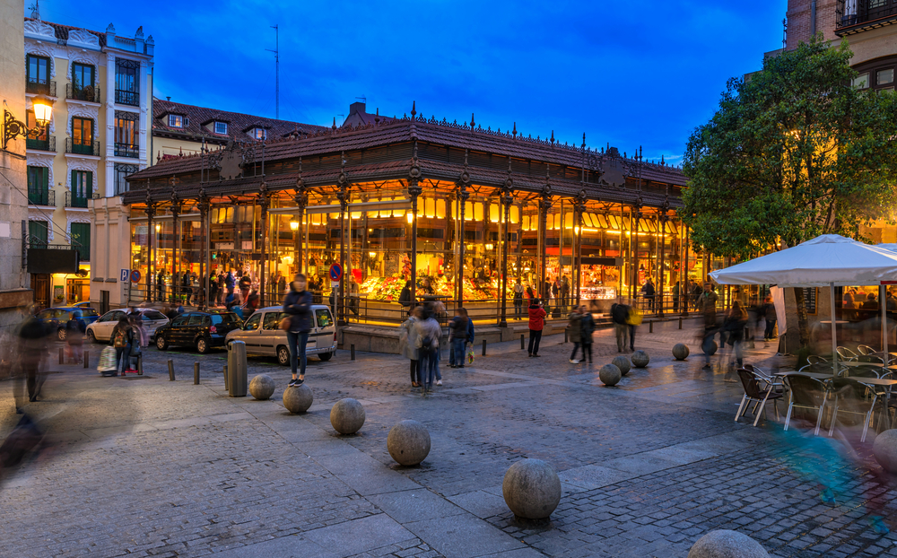 A view on a market during dusk, where various items can be seen sold inside the market, an image for a travel guide about  safety in visiting Madrid.