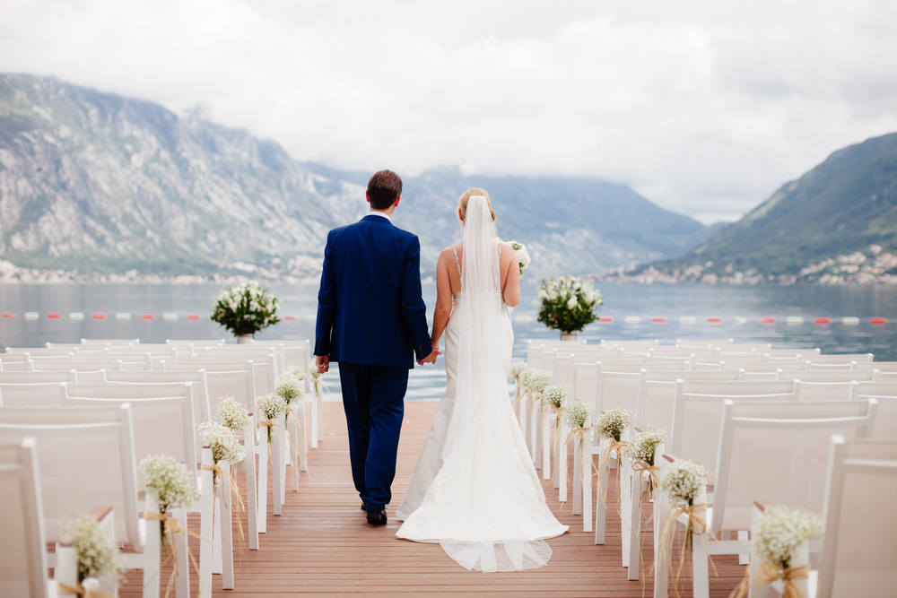 Destination wedding shown with a newlywed couple holding hands in front of mountains and the sea with empty chairs lined up