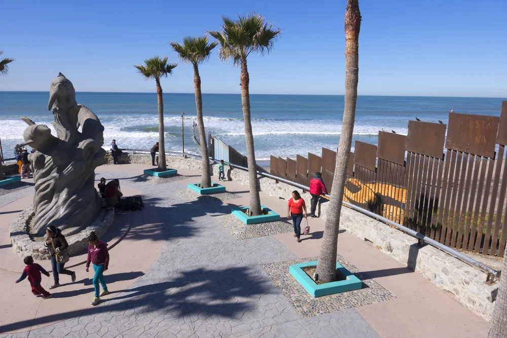People strolling around a dolphin structure near the beach with a long metal fence. 