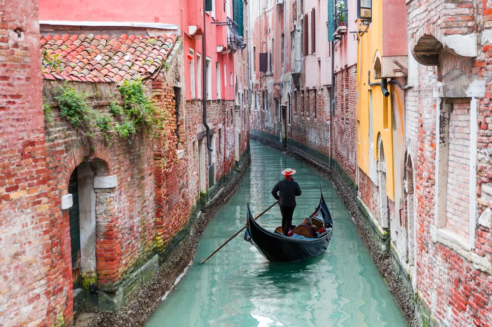 A boatman rowing his small boat in a narrow canal in between buildings. 
