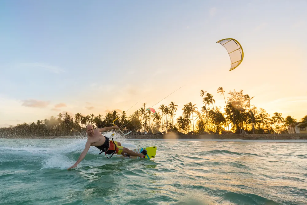 A person kiteboarding on a beach, seen gliding on the waters, and in background an island with coconut trees during sunset can be seen. 
