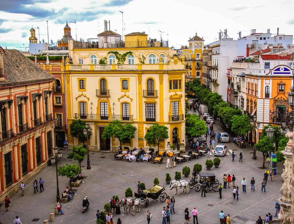 Aerial view on a historical area with a brightly painted old building in Santa Cruz, one of the best areas to stay in Seville, horse taxis parked beside the plants in the square.