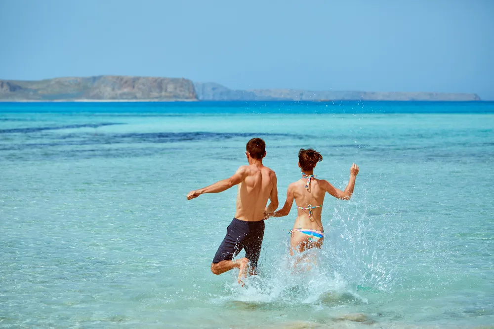 Couple excitedly running on the beach wearing swimwear while holding hands, splashing water along the way. 