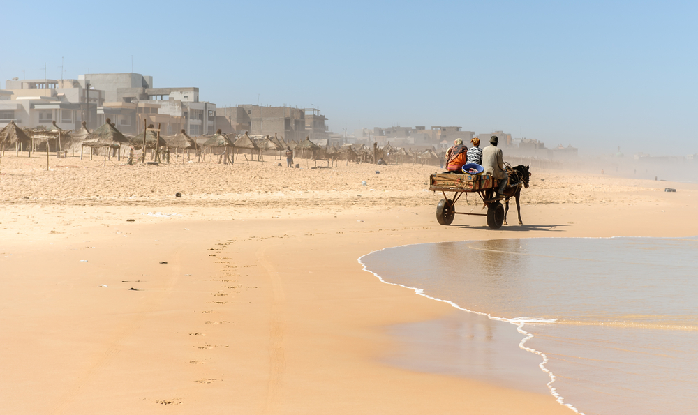 For a guide to the best and worst times to visit Senegal, a horse cart is pictured on a beach with waves gently lapping the sand and a sandstorm blowing in, seen in the distance
