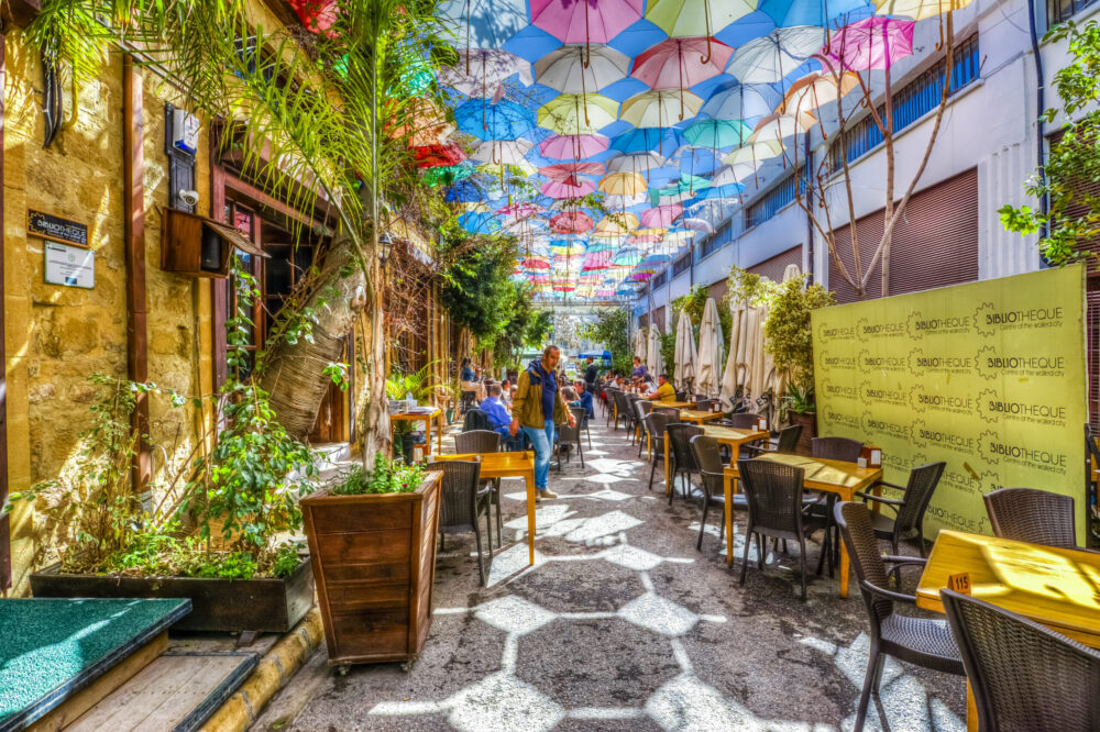 A narrow alley covered with hanging umbrellas has empty outdoor restaurant chairs and tables.