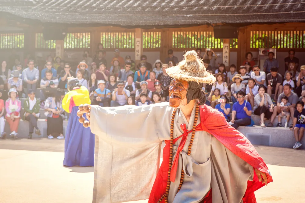 Tourists undisturbed watching a traditional asian performance where an artist is seen wearing a mask and a hay hat while dancing. 