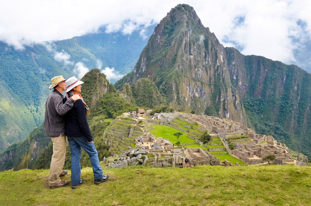 A couple wearing hats are gazing at a distance, where in background is a historical ruins and foggy mountains. 