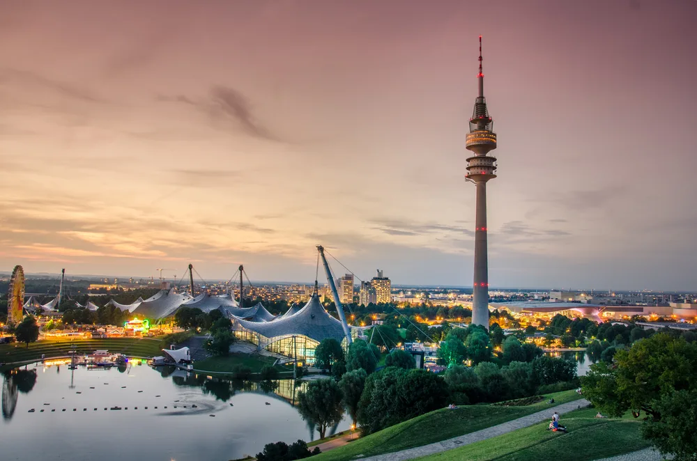 Sunset over Munich's Olympia Park, as seen at dusk with the tents being held up by crane-like devices, an image for a travel guide about safety in visiting the city.
